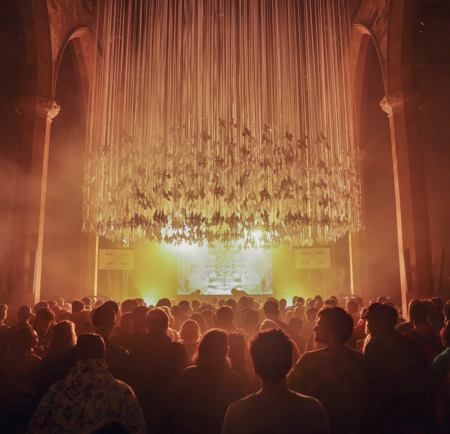 A large crowd gathered inside a grand hall with tall stone arches, facing a brightly lit stage. Above the audience hangs an elaborate installation of long, vertical ribbons or strands, illuminated by warm golden light that fills the space with a dramatic glow. The atmosphere suggests a concert or immersive art event in a historic architectural setting.