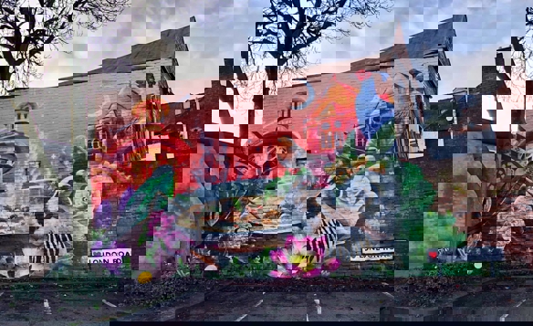 A vibrant mural painted on the side of a brick building along London Road. The artwork features a colorful nature scene with birds, flowers, and greenery, including a pheasant and a partridge near a bowl of food. Architectural elements such as domed towers and rooftops appear in the background against a red and orange sky with a crescent moon. A tree partially frames the mural on the left, and a street sign reading “♥ London Road” is visible at the bottom right.