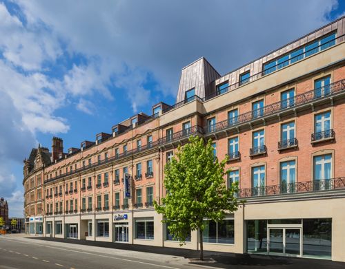 CGI-rendering of a Street view of a large, multi-story building with a red-brick façade and beige stone accents, featuring rows of tall windows with black wrought-iron balconies. The ground floor has several glass storefronts, including a visible sign for ‘Travelodge.’ A single green tree stands on the sidewalk in front of the building, and the sky above is partly cloudy with patches of blue.