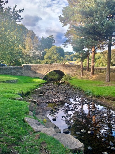 A small river filled with rocks flows towards a stone bridge. There are trees all around.