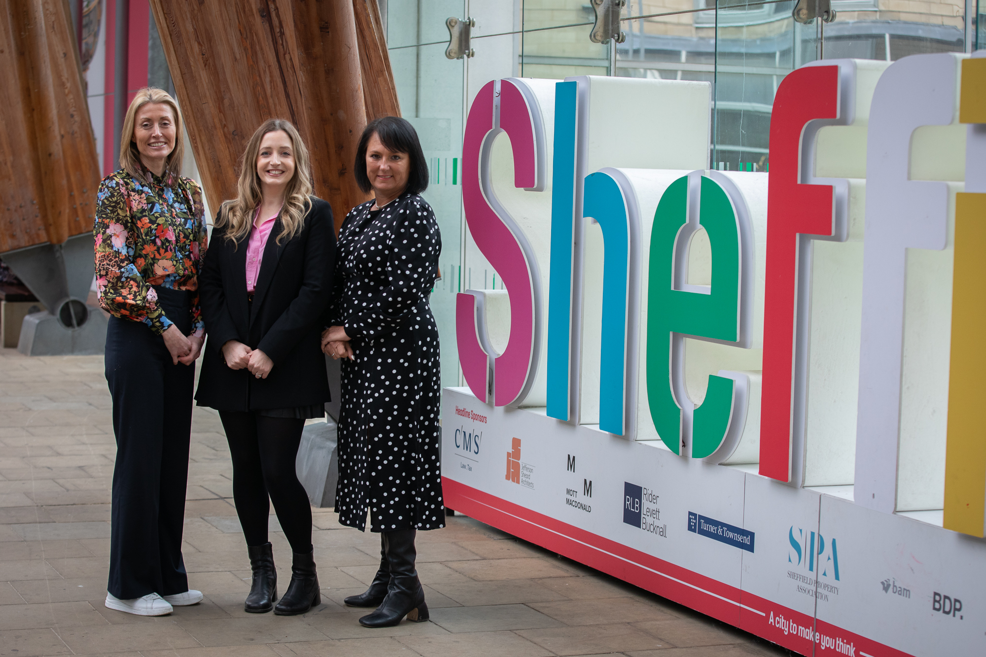 Three people standing on a paved area next to a large colorful sign that spells out part of the word “Sheffield” in bold letters. The sign features vibrant colors including pink, green, blue, and red, and is mounted on a white base with sponsor logos. Behind the group are tall wooden beams and glass panels, suggesting a modern architectural setting.