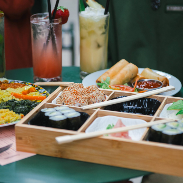 A green table filled with assorted dishes and drinks. A wooden bento-style tray holds sushi rolls, nigiri topped with avocado, sesame-coated pieces, and pickled ginger, with chopsticks resting across it. Behind the tray, a plate of golden spring rolls with dipping sauce and salad is visible, alongside a colourful platter of flatbread, shredded cabbage, and vibrant vegetable sides. Three tall cocktails garnished with fruit and herbs complete the vibrant dining scene.