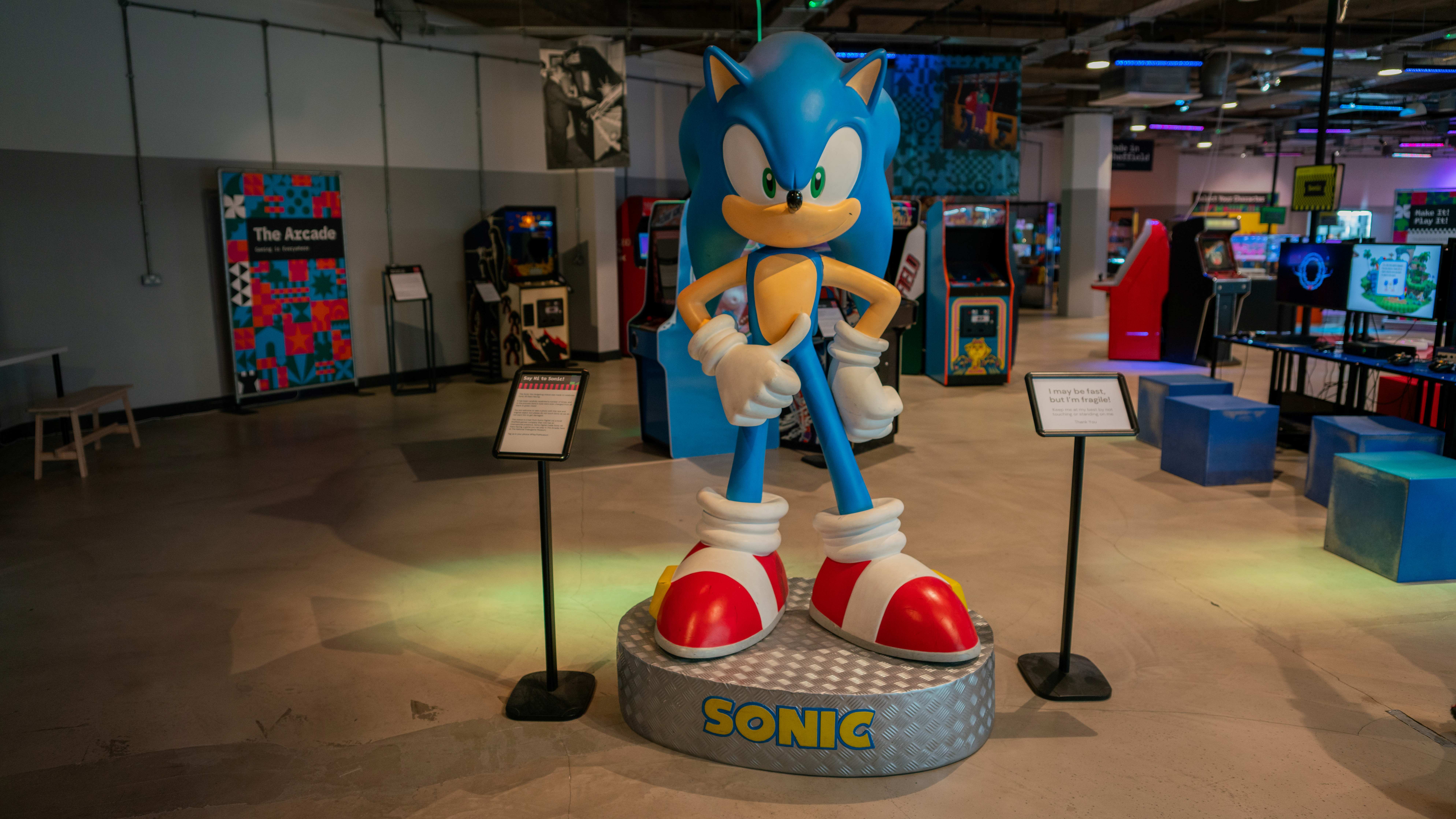 A large statue of Sonic the Hedgehog stands on a circular platform labeled “SONIC” inside the National Videogame Museum. The figure is bright blue with white gloves and red shoes, posed with arms crossed.