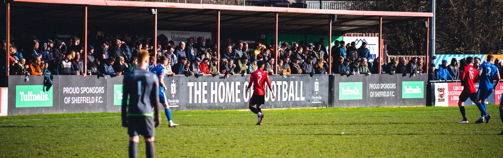 Outdoor football match at a small stadium with players on the pitch and a crowd of spectators standing under a covered stand. Advertising boards along the stand display text including ‘The Home of Football’ and sponsor logos. Trees and houses are visible in the background.