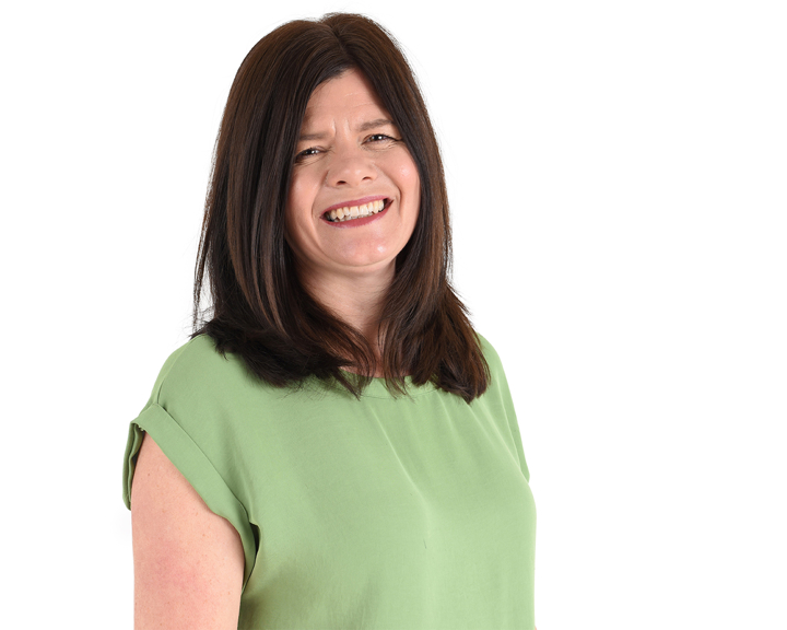 Person wearing a light green short-sleeved top, standing against a plain white background. The individual has shoulder-length dark brown hair styled straight, and the pose is slightly angled to the side.