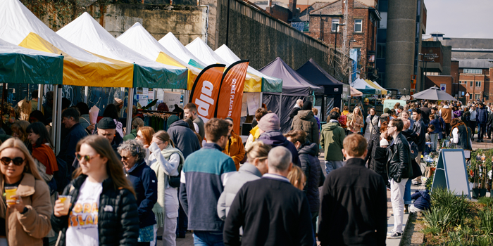 An open air market in a city centre. There are lots of people walking up and down and browsing the stalls.