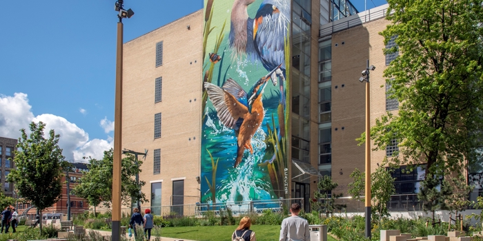 A large mural of two herons and a kingfisher in a vibrant wetland scene painted on the side of a tall modern building. In the foreground, a landscaped urban park with trees, benches, and pathways is visible, where several people, including a family with two children, are walking and enjoying the space under a bright blue sky with scattered clouds.