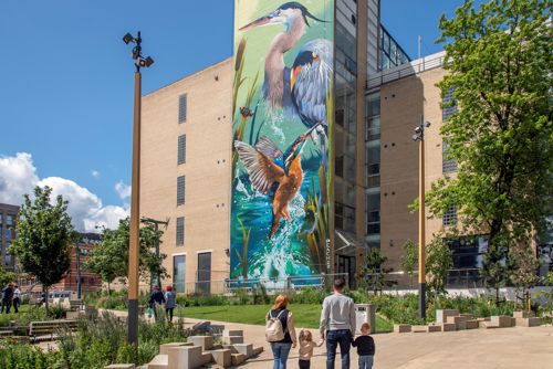 A large mural of two herons and a kingfisher in a vibrant wetland scene painted on the side of a tall modern building. In the foreground, a landscaped urban park with trees, benches, and pathways is visible, where several people, including a family with two children, are walking and enjoying the space under a bright blue sky with scattered clouds.