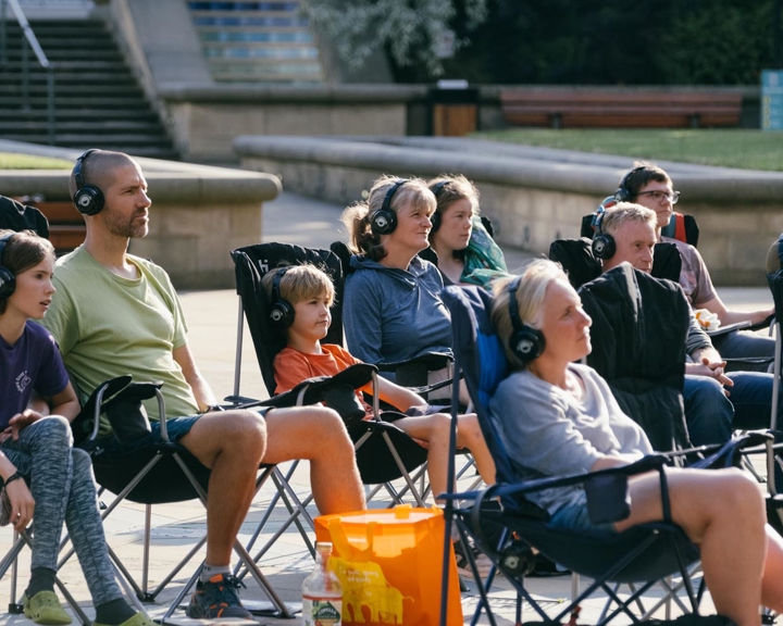 A group of people seated in folding chairs outdoors, all wearing headphones and facing the same direction as if participating in a silent event or performance. The setting appears to be a public square with stone steps, benches, and greenery in the background. A bright orange bag and a drink bottle are visible near the front row, adding colour to the scene.