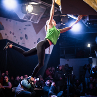Indoor climbing competition scene with a climber hanging from an overhanging wall, wearing a green top and black leggings. One arm is extended to grip a hold while the other arm reaches upward, and one leg is stretched out for balance. Bright spotlights illuminate the climber, and a crowd of spectators is visible in the background.