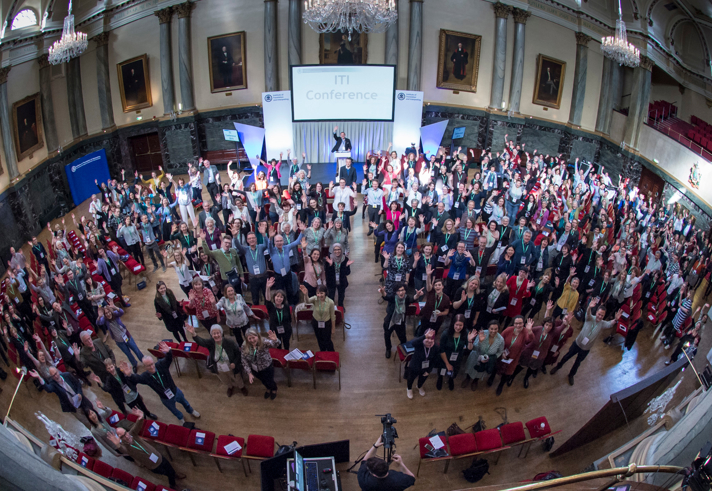 Large group of people gathered inside a grand hall with chandeliers, high ceilings, and framed portraits on the walls. The audience is standing and facing the stage, where a speaker is positioned in front of a screen displaying ‘ITI Conference.’ Rows of red chairs are visible in the foreground.