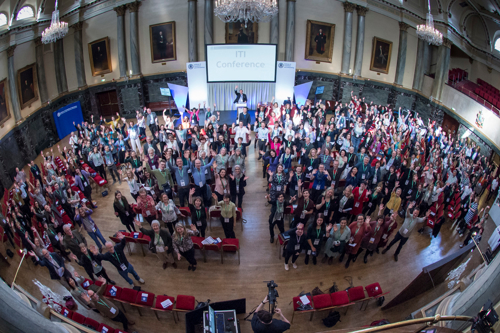 Large group of people gathered inside a grand hall with chandeliers, high ceilings, and framed portraits on the walls. The audience is standing and facing the stage, where a speaker is positioned in front of a screen displaying ‘ITI Conference.’ Rows of red chairs are visible in the foreground.