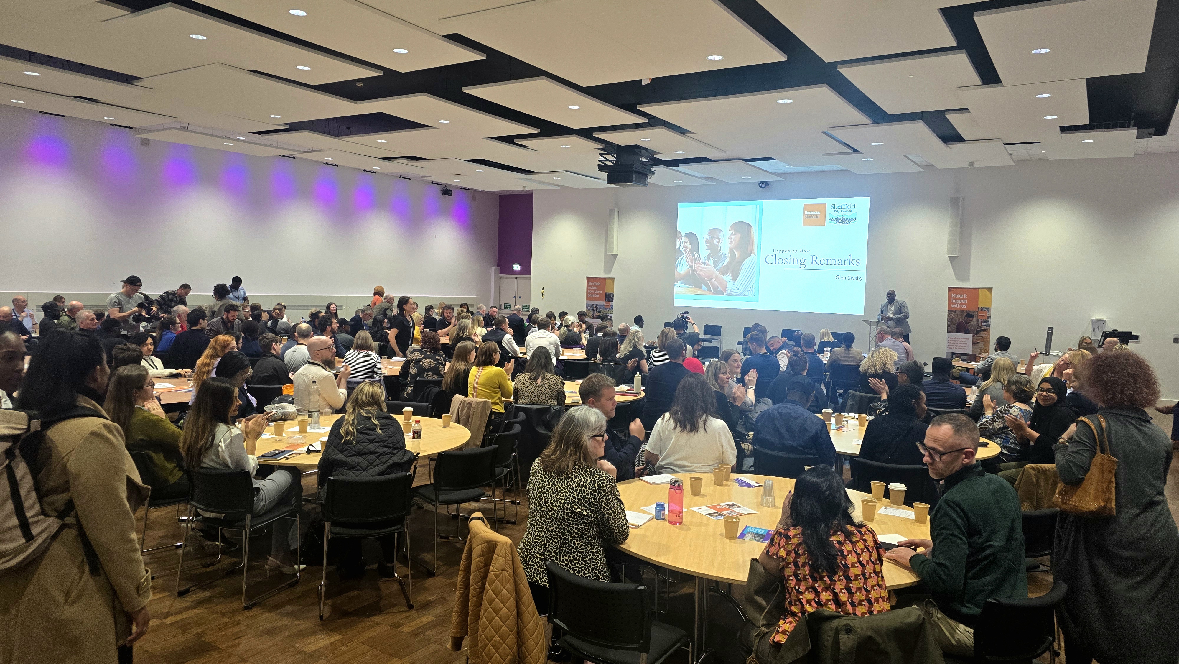 A large conference room filled with round tables where attendees are seated and engaged in discussion. The room has a modern design with a wooden floor, white walls, and a ceiling featuring black acoustic panels. Purple accent lighting runs along the left wall. At the front of the room, a large screen displays a presentation titled “Celebrating Culture & Creativity” with the James Reed logo. A speaker stands near the screen, and event banners are positioned on both sides of the stage area.