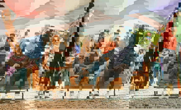 Three people sitting on an orange couch placed outdoors during a parade, with a large rainbow-colored flag held above them. The background shows a crowd of people walking and holding smaller pride flags, creating a festive and colourful atmosphere on a cobblestone street.