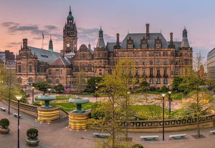 Panoramic view of Sheffield Town Hall and Peace Gardens at sunset. The historic stone building features ornate architecture with towers and a clock. In the foreground, landscaped gardens include fountains, trees, and paved walkways, surrounded by modern and traditional buildings.