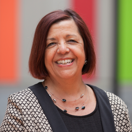 A person with short, straight auburn hair wearing a patterned jacket over a black top. The individual has a layered necklace with black oval beads. The background features vertical panels in bright colors, including red, orange, and green.
