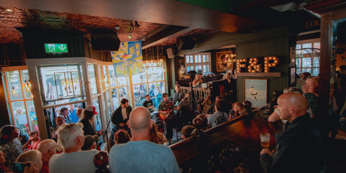 A crowded live music performance inside the Frog & Parrot pub, with warm lighting, patterned ceilings and a band playing near the front windows while people gather closely to watch.