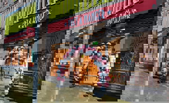 The exterior of a Pret A Manger café with a red sign displaying the name above large glass doors. A decorative arch made of maroon and white balloons stands at the entrance. The building features brick walls and bright yellow vertical panels above the storefront, and the pavement outside appears wet, suggesting recent rain.