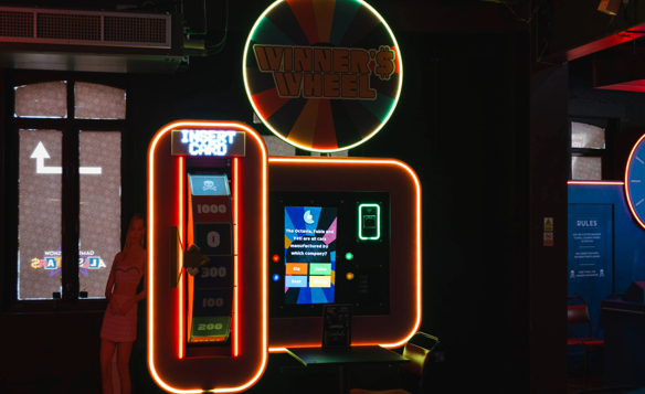 Neon-lit arcade machine called “Winner’s Wheel” with a colourful circular sign above and bright orange framing. The machine features a vertical panel with “Insert Card” text and score markers from 200 to 1000, alongside a digital screen displaying game instructions. A small table and chair sit in front of the machine, and a cardboard cut-out figure is partially visible to the left. The setting is dark with vibrant lighting, creating a lively atmosphere.