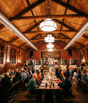 Interior of a venue with a dinner, long tables with guests sitting either side and stage at far end. 