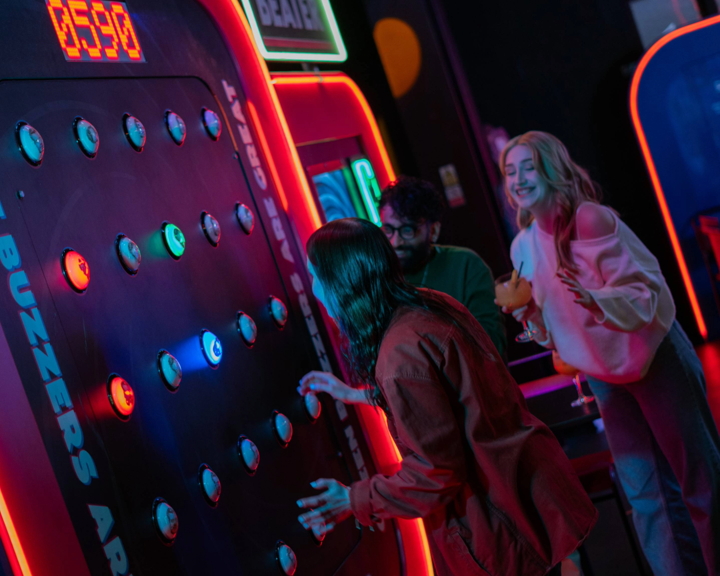 Three people at a neon-lit arcade game called “Buzzer Beater.” One person is actively playing, reaching towards illuminated buzzers on a large vertical panel displaying a score of 0590. The game features multiple round buttons glowing in blue and green, with bright signage and red lighting around the machine. Two others stand nearby, one holding a cocktail glass, watching the gameplay in a lively, colourful setting.