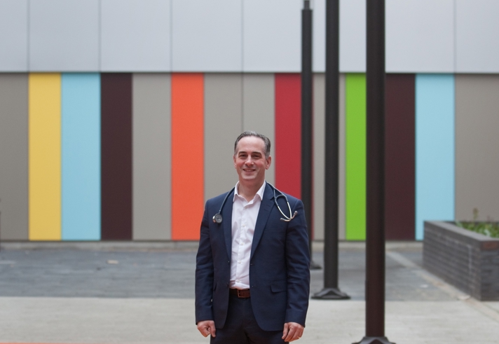 Person wearing a dark blue suit and white shirt with a stethoscope draped around the neck, standing outdoors in front of a modern building facade featuring vertical panels in bright colours including orange, yellow, green, blue, and brown. The area has a paved surface and tall black poles.