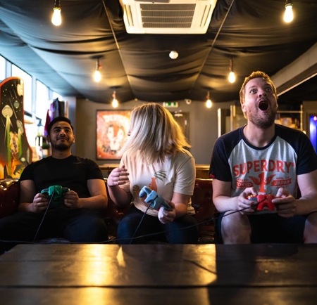 Three people sitting on a leather sofa in a gaming lounge, each holding a retro-style game controller. The room has dim lighting with exposed bulbs, arcade machines in the background, and a black ceiling with visible air conditioning unit.