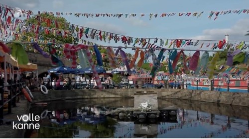 Outdoor canal-side seating area decorated with colorful fabric bunting strung across the space. People are sitting at tables under umbrellas, and narrowboats are moored along the water. The scene is bright and festive under a partly cloudy sky. Text on the bottom left reads ‘volle kanne.’