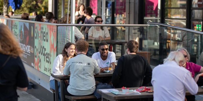 Outdoor seating area of a café or restaurant with several people sitting at wooden tables, eating and drinking. The space is enclosed by glass panels with colorful signage promoting “Sheffield DocFest” and event dates. String lights and umbrellas provide shade, and the background shows more tables and people inside the establishment.