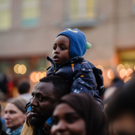 A child wearing a blue hat and padded jacket sits on someone’s shoulders in a crowded outdoor setting. The background shows blurred lights and a building, suggesting an event or gathering.