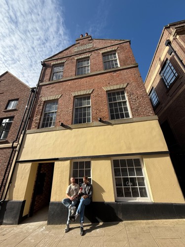 Tall brick building with three stories and a cream-colored ground floor facade. The upper floors have large rectangular windows with stone lintels, and the roofline features a small decorative gable with engraved text. Two people are sitting on the low ledge in front of the building, near a window with the word ‘Güs’ visible on the glass. The scene is lit by bright sunlight under a clear blue sky with wispy clouds.