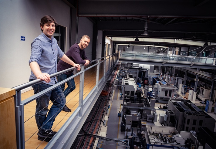 Two people standing on an upper-level walkway inside a large industrial facility, leaning on a metal railing and overlooking rows of advanced manufacturing machines and equipment below. The space features high ceilings, exposed beams, and bright overhead lighting, creating a modern factory environment.
