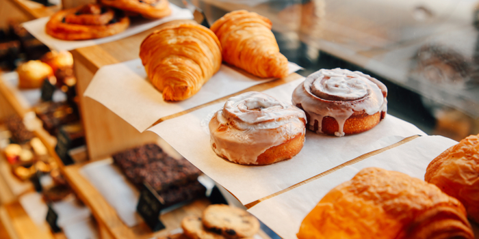 Bakery display with wooden shelves holding a variety of pastries. Visible items include golden croissants, iced cinnamon rolls, pain au chocolat, chocolate brownies, and cookies arranged on trays lined with parchment paper. Sunlight streams through the window, illuminating the baked goods.
