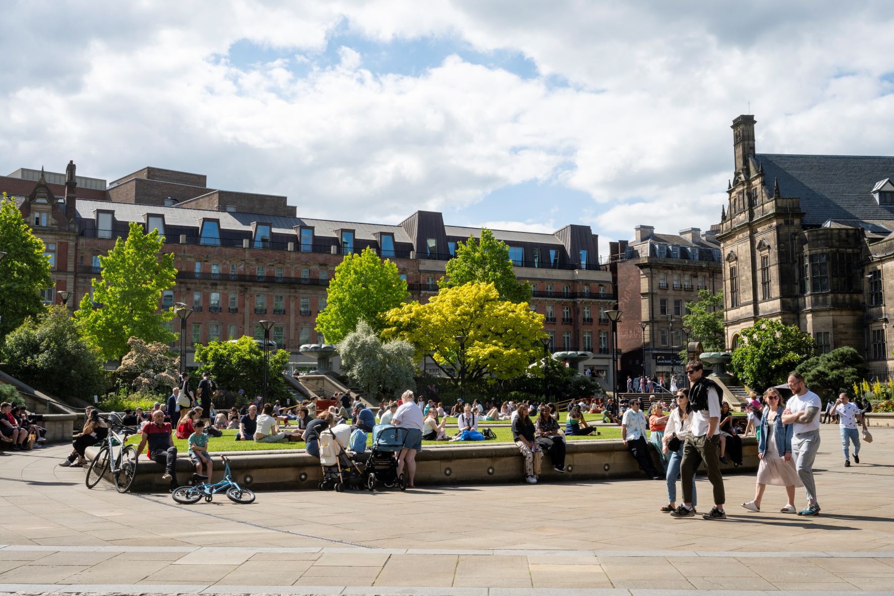 Lots of people enjoying a warm sunny day in The Peace Gardens. In the background is The Radison Blu Hotel.