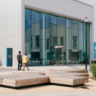 Exterior view of a modern building with large glass panels and a white, translucent facade. Two people walk toward the entrance, which features a glass door. In front of the building, there are geometric wooden benches on a paved area, with a blue signboard visible on the left side.