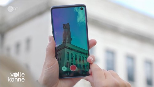 Close-up of a smartphone held in both hands, showing a live camera view of a building with a large sculpture of a cat on its roof. The phone screen displays a red record button, and the background shows the same building slightly out of focus. Text on the bottom left reads ‘volle kanne.’