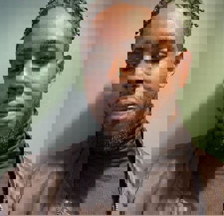 Person wearing a dark turtleneck sweater and a black jacket, standing against a plain green wall. A small round fixture is visible on the wall near the top right of the image.