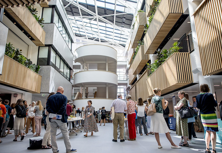 Spacious modern atrium with a glass ceiling and natural light, featuring multiple floors with wooden slatted balconies and green plants. A group of people is gathered in the open central area, some standing near tables with refreshments. The design includes curved white staircases and sleek architectural details, creating a bright and contemporary setting.