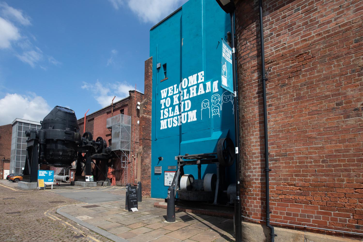 The entrance to the Kelham Island Museum. On the right is a blue-painted wall with 'Welcome To Kelham Island Museum' on it. In the background you can see a huge Bessemer Converter that was used in the production of steel.