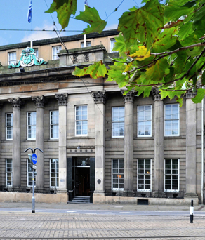 The exterior of Cutlers' Hall. A historic stone building with neoclassical architecture featuring tall columns, large rectangular windows, and ornate detailing above the entrance. The facade includes decorative carvings and a crest at the top center. Tram tracks run along the street in front, and green leaves from a nearby tree frame the upper part of the image.