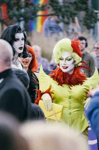 Two individuals in elaborate costumes pose outdoors. One wears a black and white outfit with long black hair and white face makeup. The other has bright green hair, dramatic makeup, and a vibrant green outfit with red feather accents, holding a yellow clutch. Rainbow flags and onlookers are visible in the background.