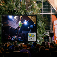 A large indoor screen displays a climber in a green top hanging from a bouldering wall during the CWIF 2024 event. The screen is set up in a spacious venue filled with lush green plants and wooden beams, with rows of seated spectators watching. An orange banner with text stands to the right, and bright lighting highlights the modern glass structure in the background.