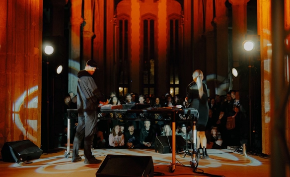 Two performers stand behind a DJ table in a grand hall with tall columns and arched windows, illuminated by warm orange lighting and spotlights. A seated audience watches in the background.