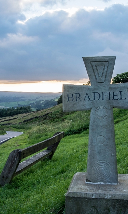 On a grass verge between two roads stands a stone cross with the word 'Bradfield' carved into it.