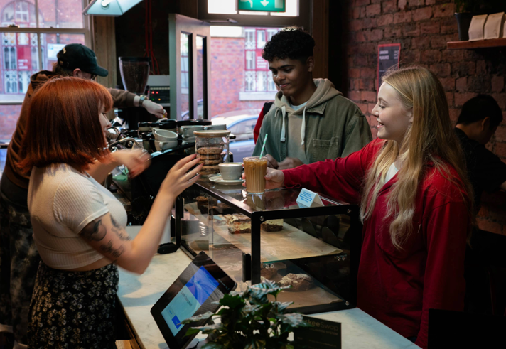 A barista is serving drinks at a coffee shop counter with a glass display case containing pastries. Two customers stand on the other side of the counter, one holding a takeaway cup with a straw. Behind the counter, coffee-making equipment and jars of biscuits are visible. The setting features exposed brick walls, a plant on the counter, and a window showing a red-brick building outside.