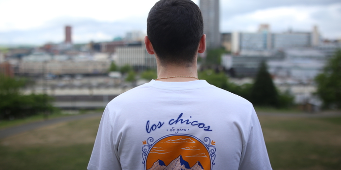  Person wearing a white T-shirt with a colorful back design showing mountains, a winding river, and an orange sunset. The text reads “los chicos de gira” in blue script. The background features a cityscape with tall buildings and greenery.