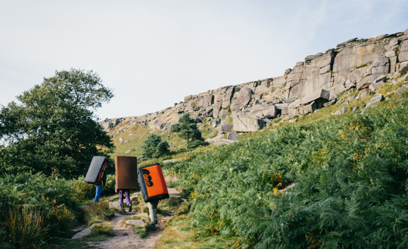 Three people are walking along a path that leads to a huge gritstone rock face. They are all carrying climbing crashmats on their backs.