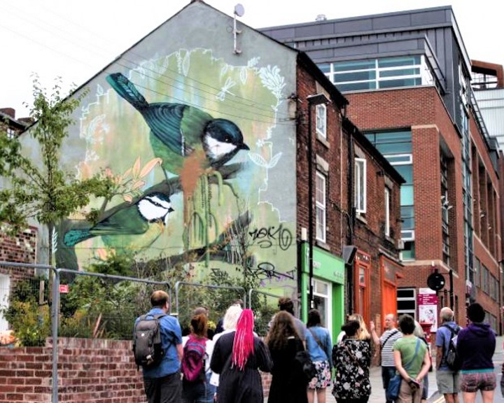 Group of people walking down an urban street toward a building with a large mural of two birds perched on branches, painted on the side wall. The mural features natural tones of green and brown, and the surrounding area includes brick buildings, a green shopfront, and modern architecture in the background.
