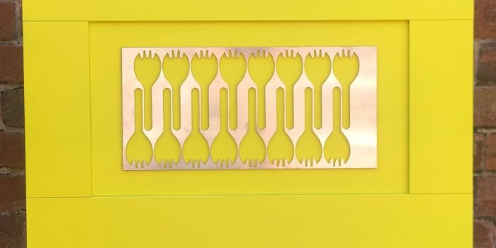 Contemporary artwork displayed on an easel, featuring a bright yellow frame and a central metallic panel with cut-out fork-like shapes, set against a brick wall background.