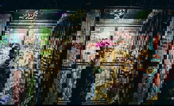 Person standing inside a graffiti-covered glass shelter at night, surrounded by colourful tags and street art illuminated by moody lighting.